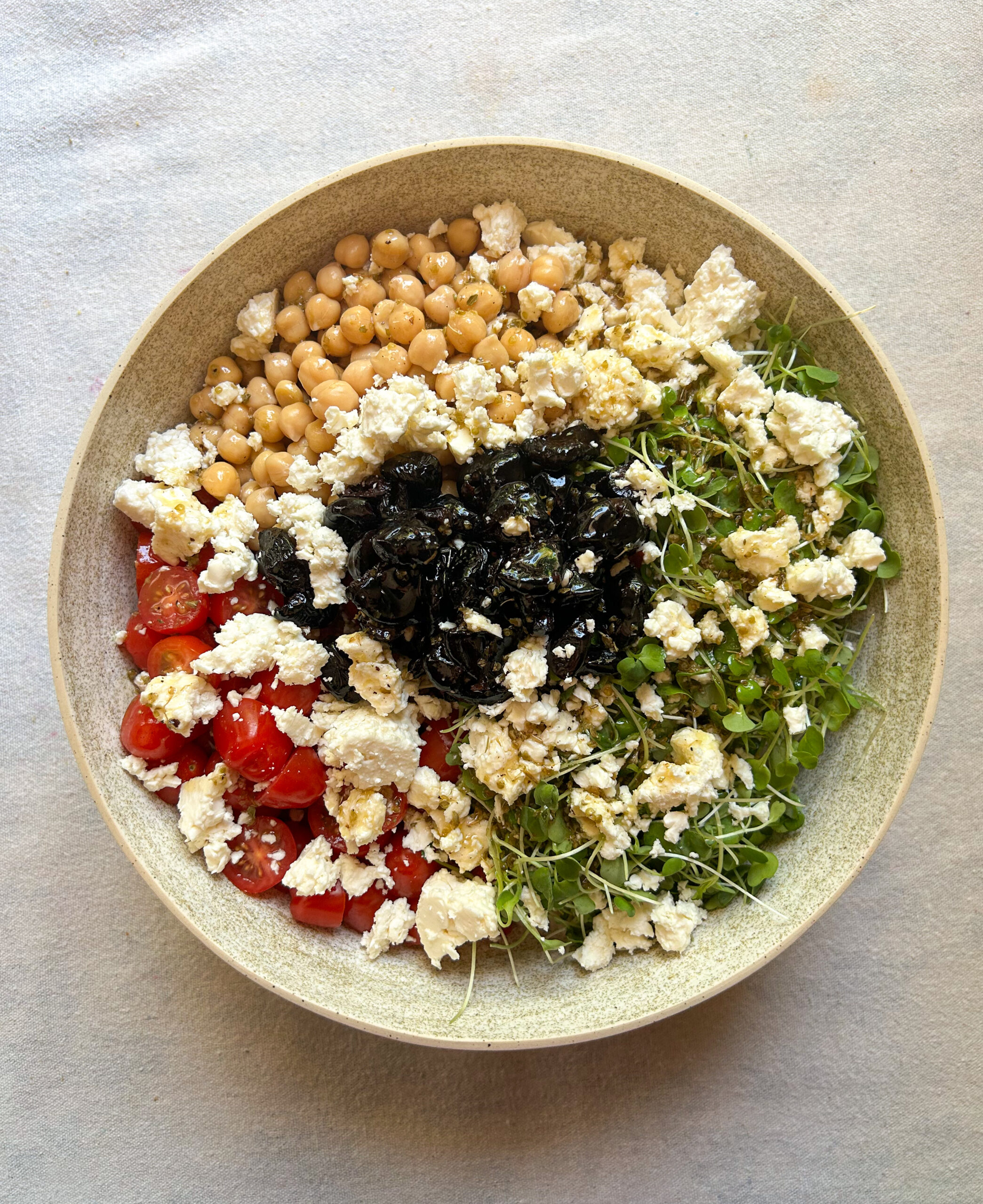 A large bowl sits on a table filled with couscous, arugula, tomatoes, chickpeas, olives and feta.