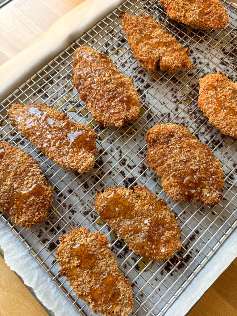 baked chicken tenders covered in a drizzle of hot honey and a sprinkle of flakey salt sit on a wire rack-lined baking sheet on a wooden counter
