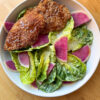 oven baked chicken tenders on a bed of baby gem salad in a white bowl sits on a wooden counter top