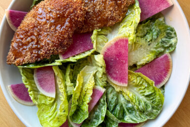 oven baked chicken tenders on a bed of baby gem salad in a white bowl sits on a wooden counter top