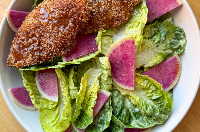 oven baked chicken tenders on a bed of baby gem salad in a white bowl sits on a wooden counter top