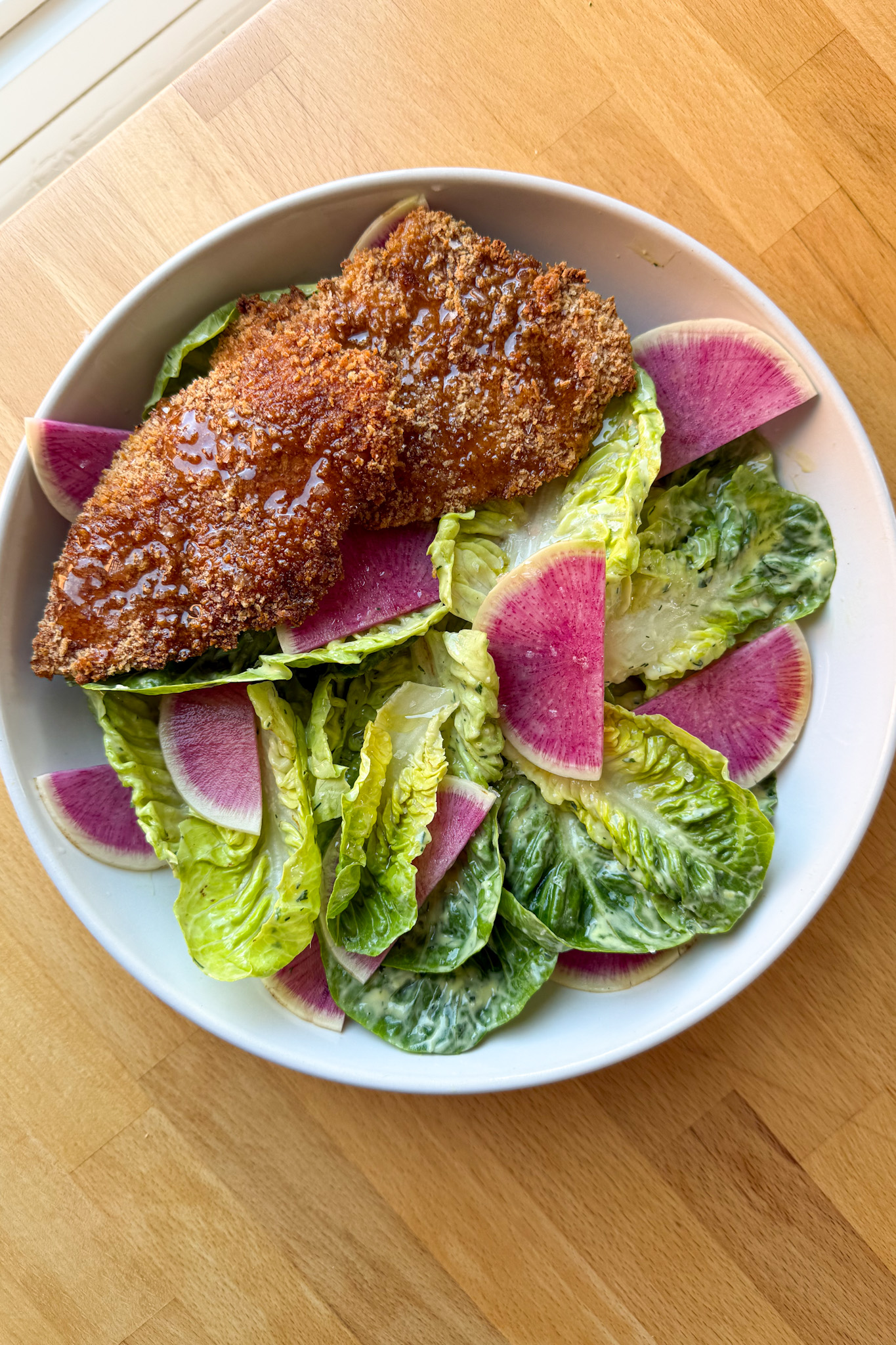 oven baked chicken tenders on a bed of baby gem salad in a white bowl sits on a wooden counter top