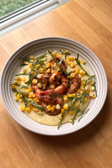 a white plate sits on a wooden counter filled with creamy corn polenta, seared cajun shrimp and fresh basil