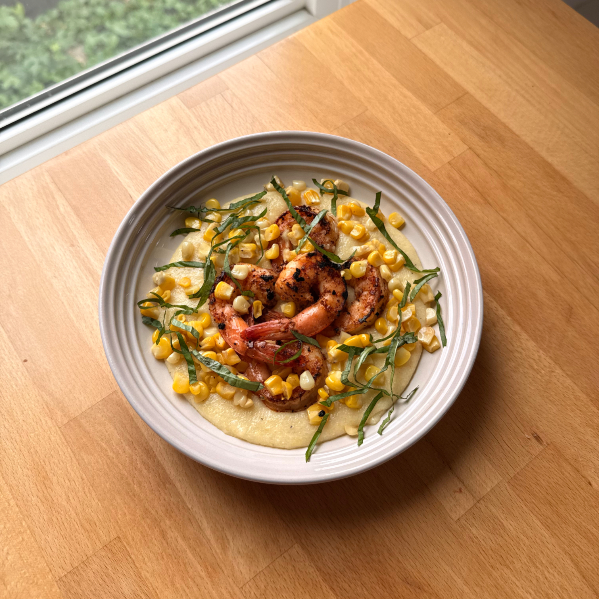 a white plate sits on a wooden counter filled with creamy corn polenta, seared cajun shrimp and fresh basil