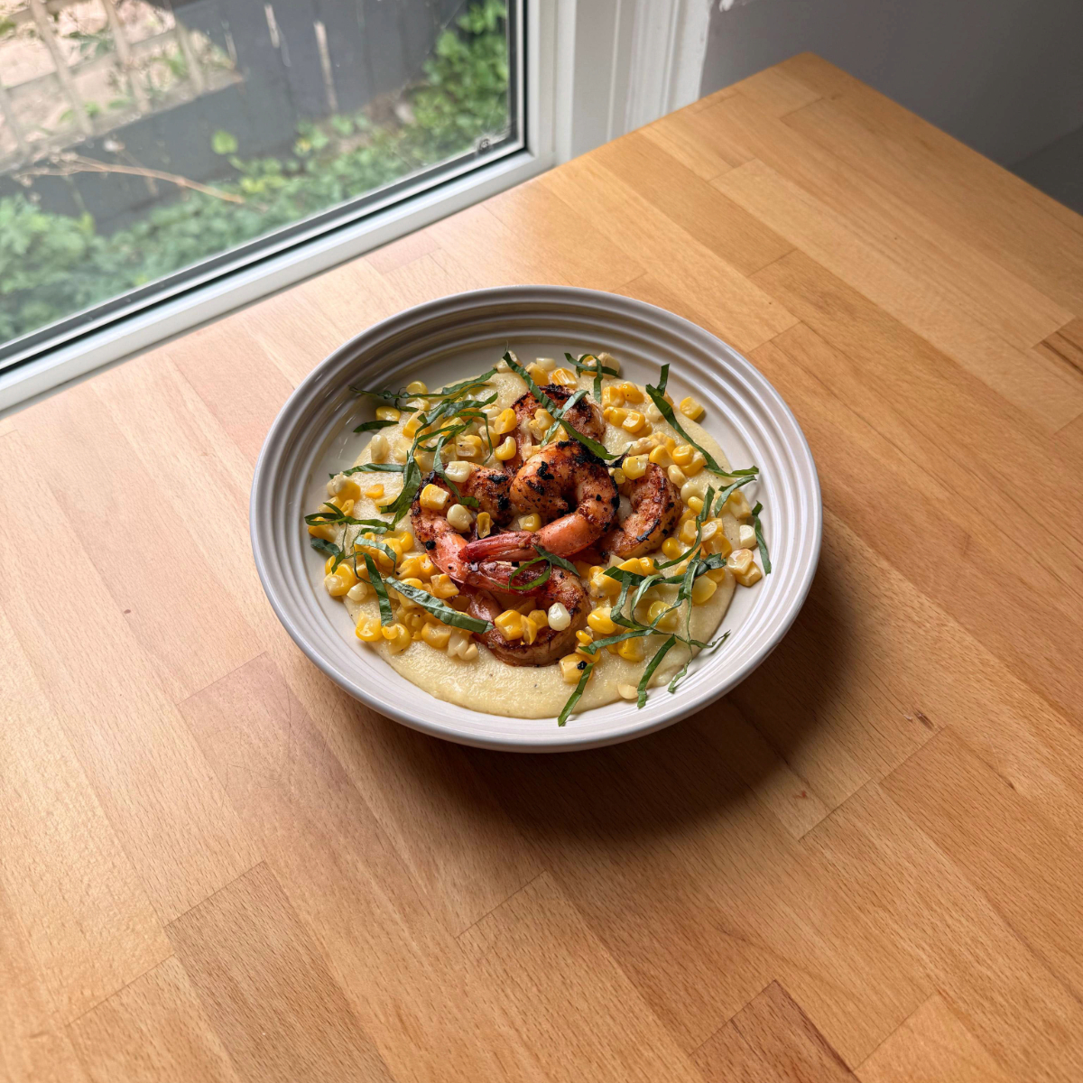 a white plate sits on a wooden counter filled with creamy corn polenta, seared cajun shrimp and fresh basil