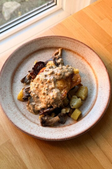 an oven baked chicken thigh on a plate with potatoes and mushrooms topped with au poivre sauce